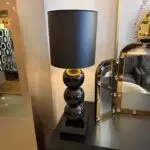 Glossy black table lamp with stacked orb base and cylindrical shade on a black square pedestal in a showroom setting.