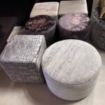 Group of silver-gray upholstered ottomans and stools arranged together, including round floral-patterned tops and square geometric-patterned tops.