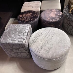 Group of silver-gray upholstered ottomans and stools arranged together, including round floral-patterned tops and square geometric-patterned tops.