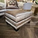 Luxurious showroom scene with brown velvet tufted chairs and matching ottoman on a dark wood floor, beige sofa in background.