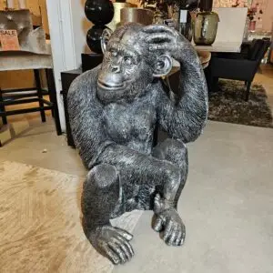 Gray gorilla statue seated with one hand resting on its head inside a showroom surrounded by furniture and decor.
