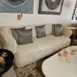 Beige sofa with black-and-white geometric pillows in a showroom; sale tags and glassware on a nearby white table in front.