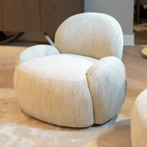 Close-up of a light gray, rounded upholstered armchair on a beige carpet in a modern living room. Alternative: Soft textured curved armchair with neutral tones in an upscale living area.