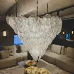 Close-up of a decorative glass chandelier hanging over a beige living room sofa area, with fruit bowls and pale flowers on a coffee table in the foreground.