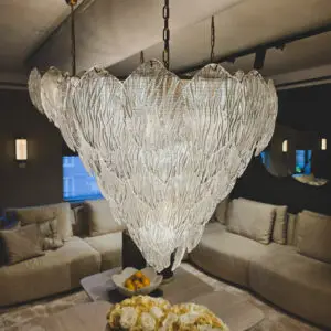 Close-up of a decorative glass chandelier hanging over a beige living room sofa area, with fruit bowls and pale flowers on a coffee table in the foreground.