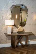 Curved console table with a wavy-edge mirror, lamp, crystal cluster, and stacked books against a textured tiled wall.