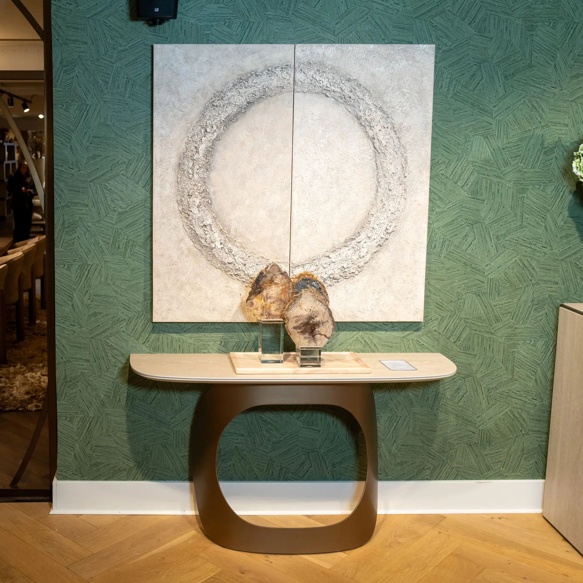 Two-panel stone artwork with a circular textured ring mounted on a green patterned wall, placed above a curved brown pedestal table with small rock sculptures in front.