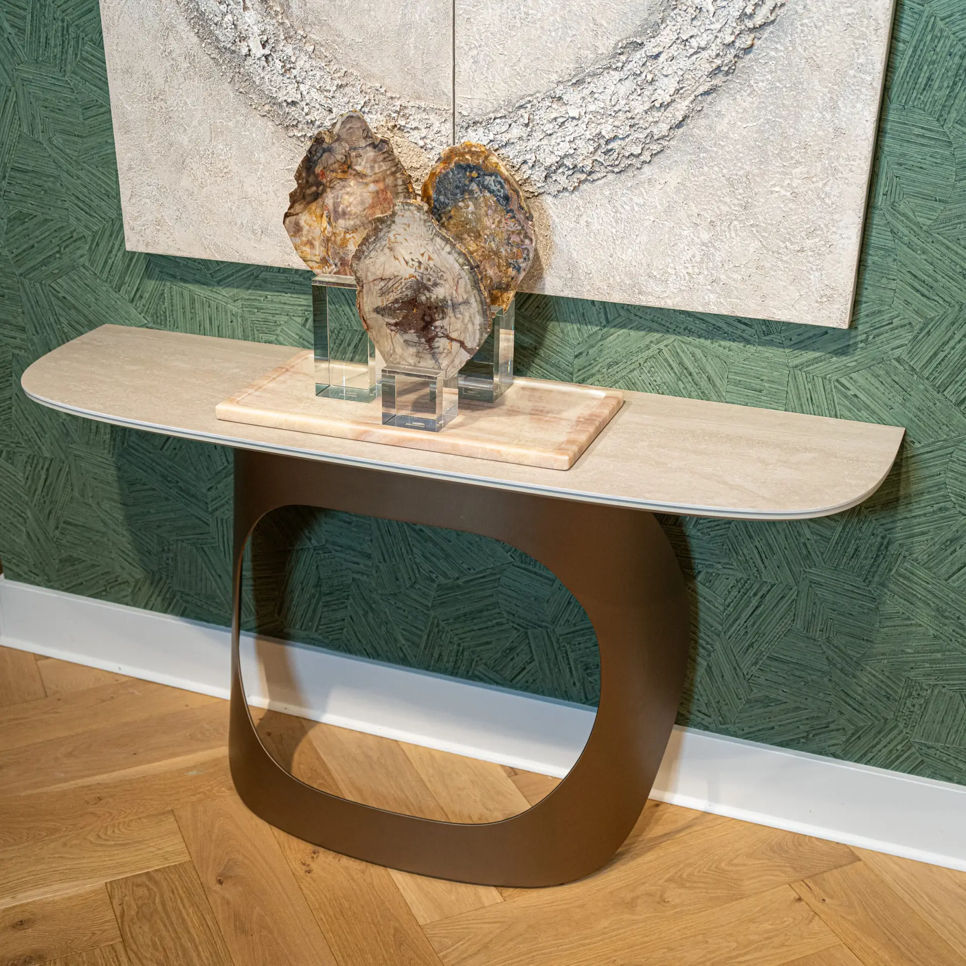 Display view of a sculptural brown curved chair with glass supports in front of a textured stone panel artwork on a green patterned wall, atop a wooden floor.