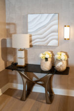 Framed abstract artwork with white and beige wavy patterns, hung on a textured gray wall, beside a beige lamp and two vases of white roses on a dark console table.