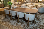 Elegant living-droom showroom with a wooden dining table and light gray upholstered chairs on a shaggy rug, greenery centerpiece in focus.
