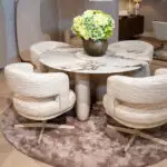 Marble round dining table with a green hydrangea bouquet in a dark ribbed vase, surrounded by textured beige chairs in a showroom.