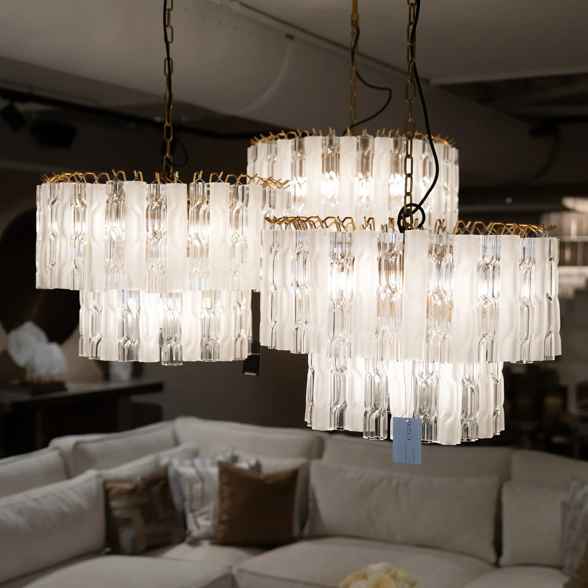 Elegant glass chandelier with multiple cylindrical glass shades suspended from gold chains in a furniture showroom with beige sofas in the background.