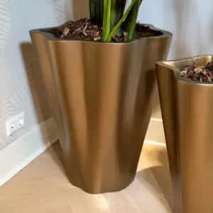 Tall bronze planter with green plant stems and brown mulch indoors, near a wall outlet and light wood floor.