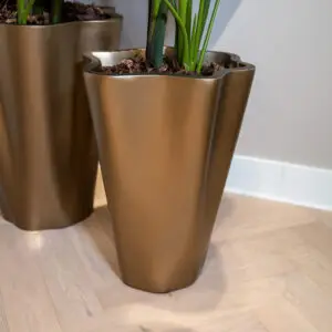 Close-up of a tall bronze planter with a green plant stem, on a light wood floor; a second planter is blurred in the background.