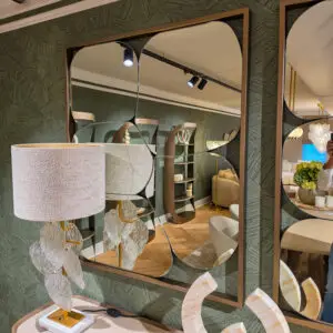 Interior of a home decor showroom with rounded rectangular mirrors in bronze frames mounted on a green geometric wallpapered wall, reflecting track lighting and furniture.