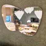 Two overlapping round mirrors with copper frames reflect a modern living room: a beige sofa, a chandelier, and potted plant against a textured wall.