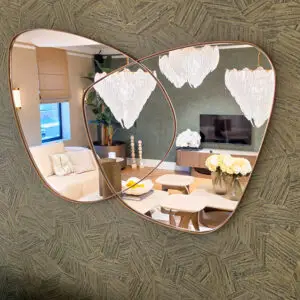 Two overlapping round mirrors with copper frames reflect a modern living room: a beige sofa, a chandelier, and potted plant against a textured wall.