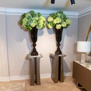 Two tall dark vases holding large green hydrangea bouquets mounted on a wall, with small pedestal tables beneath and track lighting above.