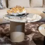 Crystal cluster or quartz geode resting on a closed book atop a marble coffee table in a living room setting, with a blurred sofa in the background.