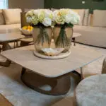 Two white rose bouquets in textured glass vases sit on a round coffee table in a cozy living room.