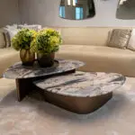 A round marble coffee table with three stacked surfaces holds two vases of green hydrangeas in a living room with a beige sofa and textured pillows in the background.