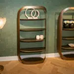 Modern display shelf against a teal geometric-patterned wall, featuring two circular stone sculptures on the top shelf and white roses in vases on the right.