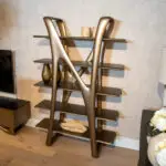Metallic sculptural display stand with three shelves, holding two stones, placed against a patterned wall backdrop.