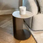 White candle in a white ceramic jar with a small gold square logo sits on a round brown side table next to a beige sofa in a living room.
