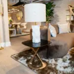 A modern living room scene with a beige lamp on a white pedestal table, brown sofa, and patterned cushions on a shaggy rug.
