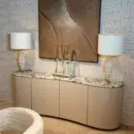 Bedroom vignette: curved beige dresser with marble top, two white textured lamps, and glass sculpture accents in front of abstract patterned walls.