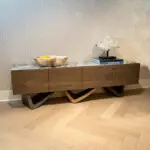 Wooden sideboard with a marble bowl on the left and a crystal display on a stand with stacked books on the right, against a geometric wallpapered wall.