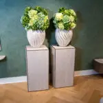Two beige textured vases with large green hydrangea arrangements on wooden display plinths against a teal patterned wall.