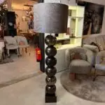 Tall glossy black stacked-sphere floor lamp with a large textured lampshade, in a furniture showroom surrounded by gray upholstered chairs and display shelves with sale tags.