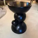 Tall black glossy sculpture made of stacked spheres with a large shallow bowl on top, standing on a rounded base in an indoor setting.