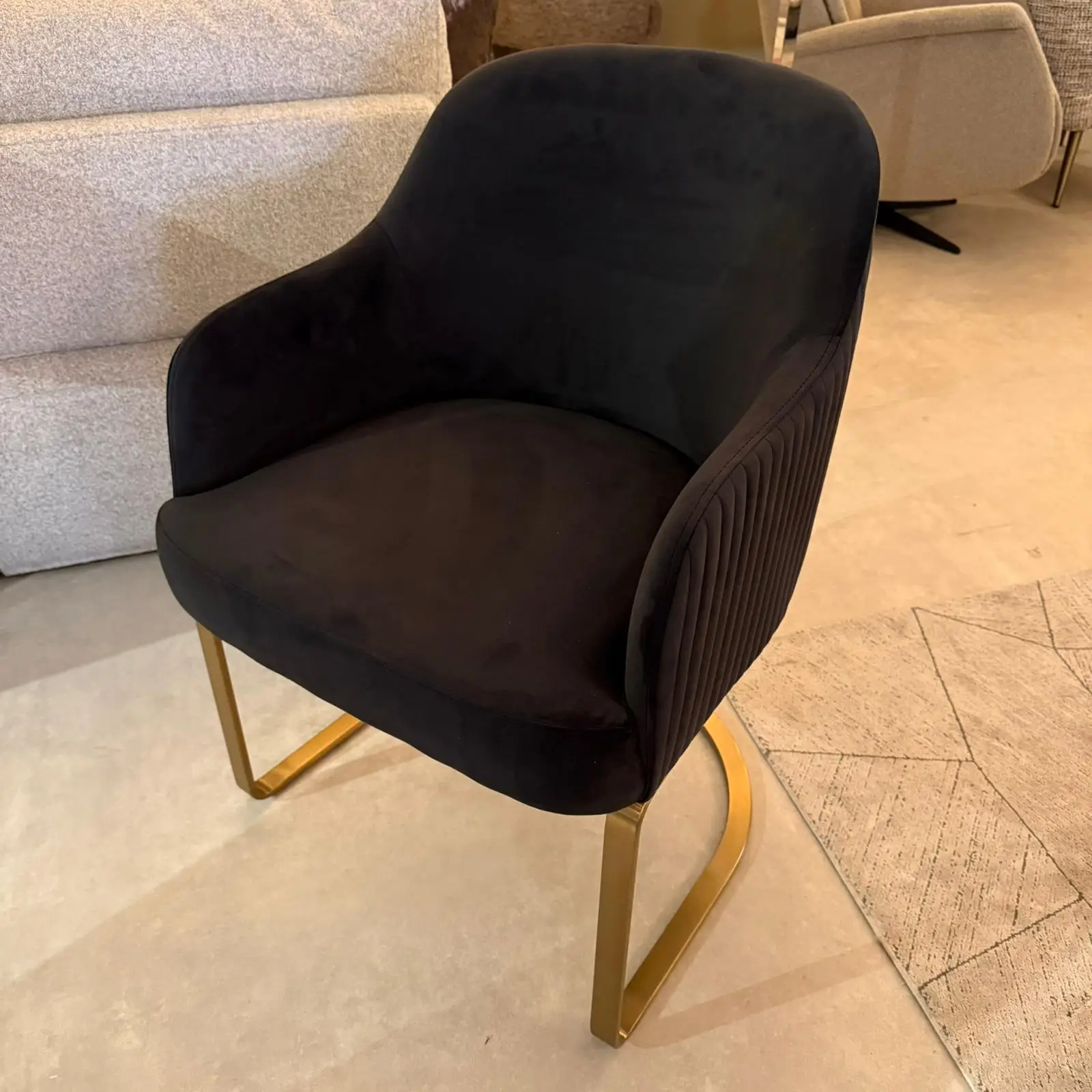 Black upholstered armchair with curved back and gold-tone metal legs in a showroom setting, surrounded by other furniture pieces.