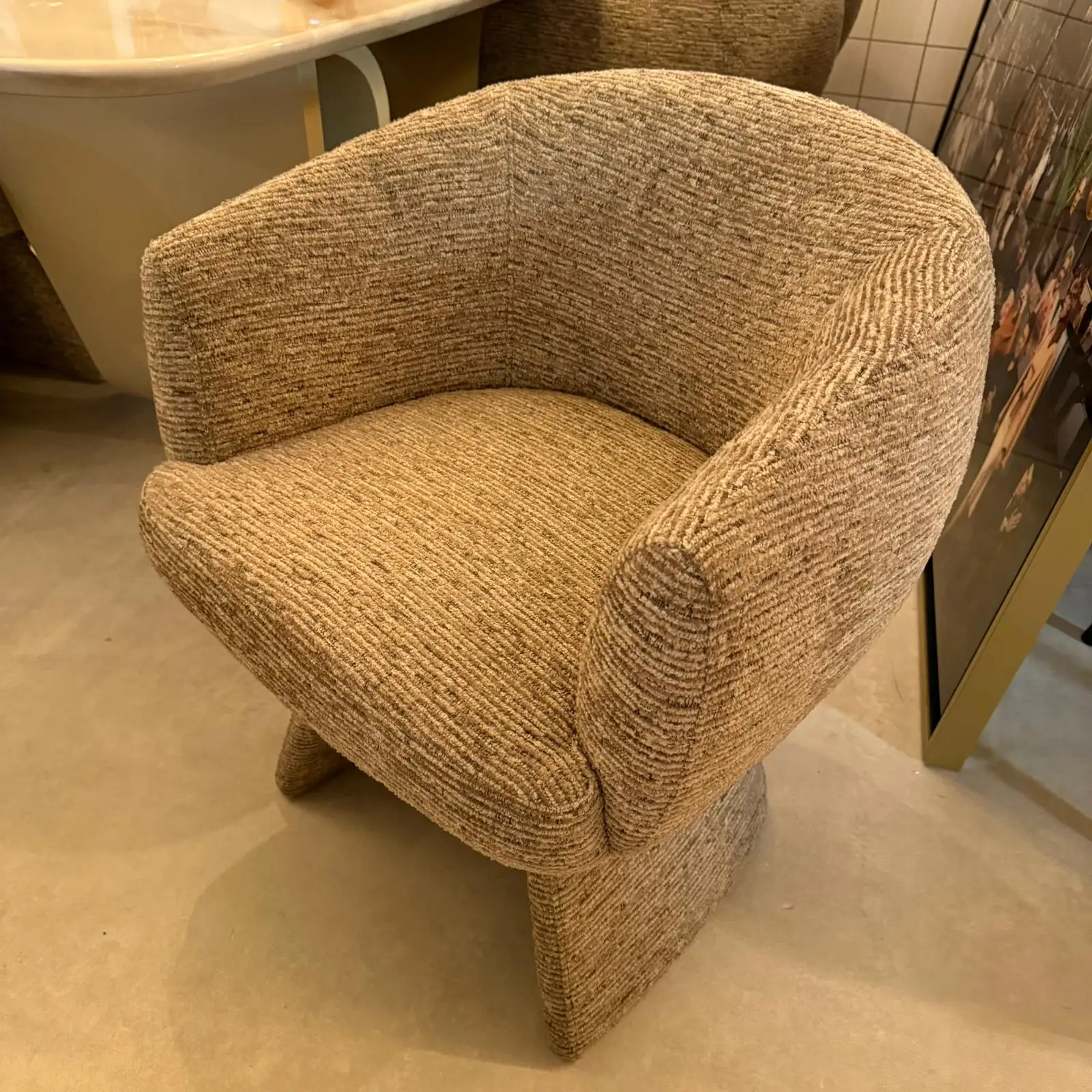 Beige textured upholstered armchair with rounded arms, shown in a showroom setting next to a sink and display cabinet.
