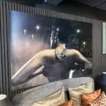 Large wall art showing a woman's back with an ornate diamond necklace, mounted on a dark striped wall above a gray sofa with tan cushions.