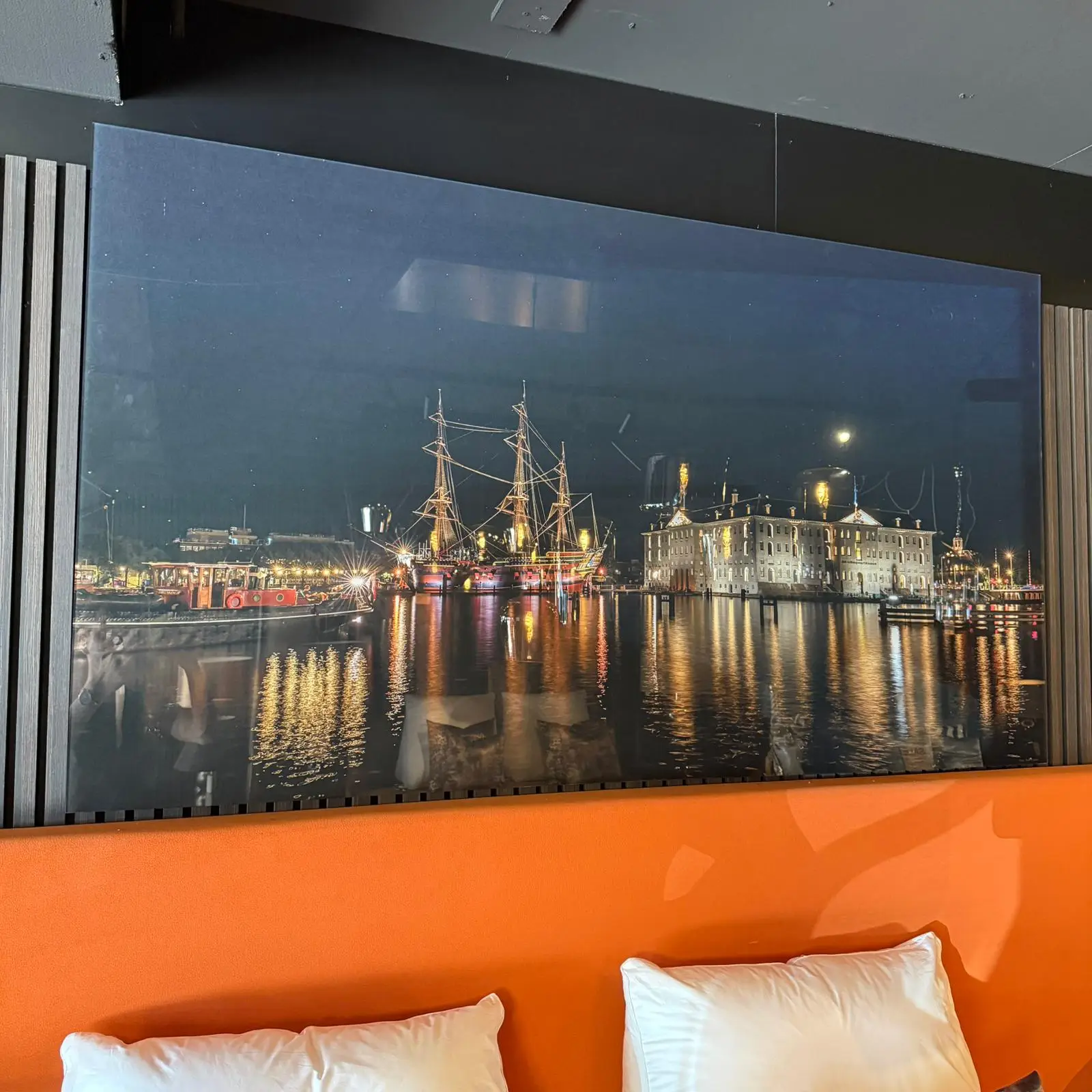 Nighttime harbor scene depicted as wall art: a lit ship beside a white-brick building reflected in calm water, hung above an orange wall with white pillows nearby.
