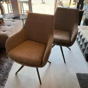 Two brown upholstered lounge chairs with metal legs in a showroom, beside a tufted ottoman and glass tables near large windows.