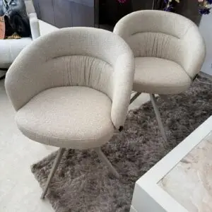 Two beige curved armchairs with pleated backs on a gray shag rug in a showroom setting.