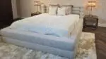 Gray upholstered platform bed with a pale quilted cover and white decorative pillows, flanked by brass lamps on matching nightstands against a textured wallpaper backdrop.