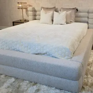 Gray upholstered platform bed with a pale quilted cover and white decorative pillows, flanked by brass lamps on matching nightstands against a textured wallpaper backdrop.
