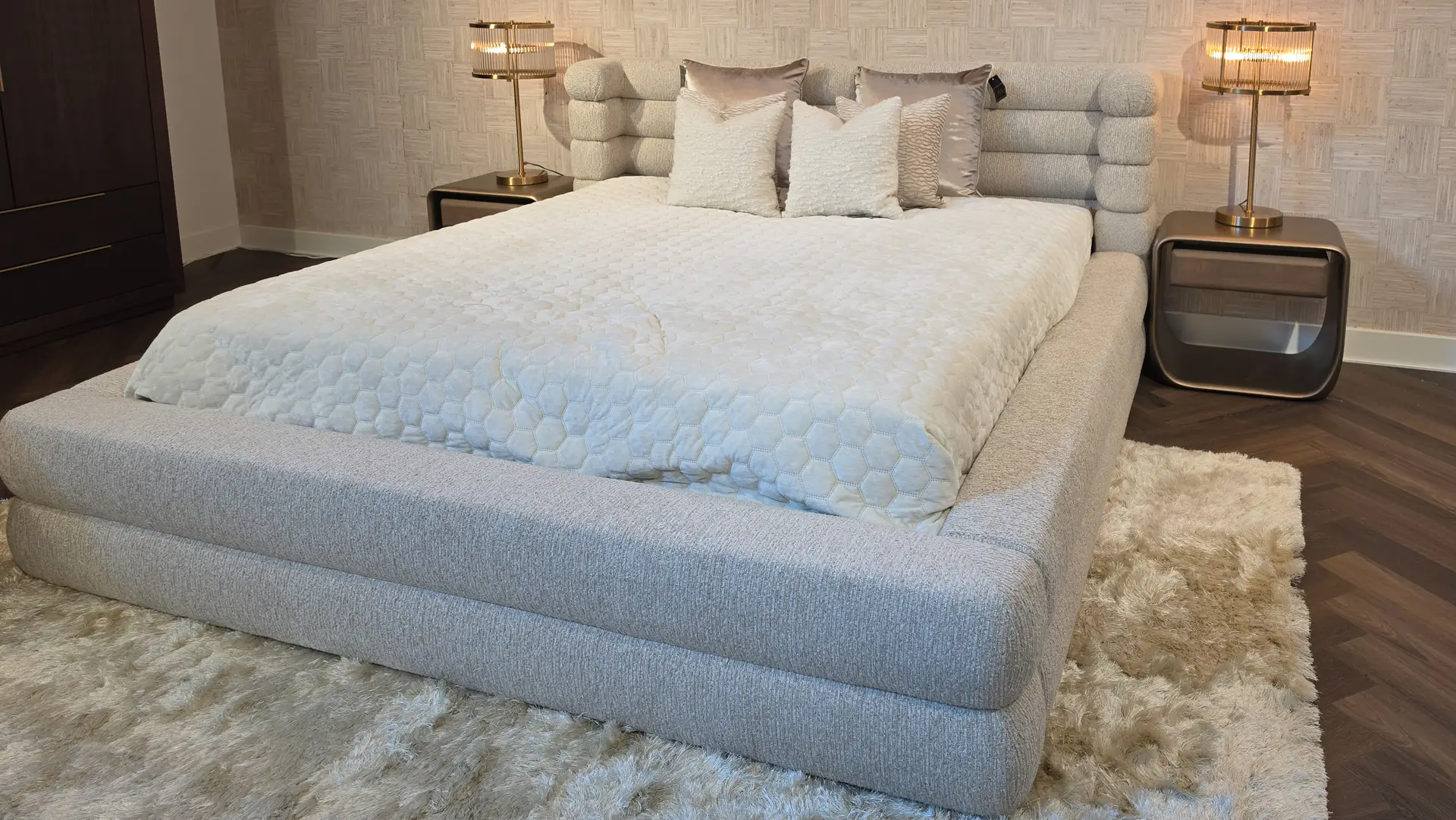 Gray upholstered platform bed with a pale quilted cover and white decorative pillows, flanked by brass lamps on matching nightstands against a textured wallpaper backdrop.