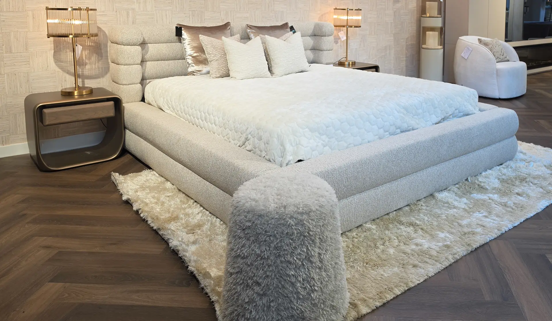 Luxurious light-gray upholstered bed with quilted bedding and multiple pillows in a showroom setting, flanked by matching nightstands and brass lamps.