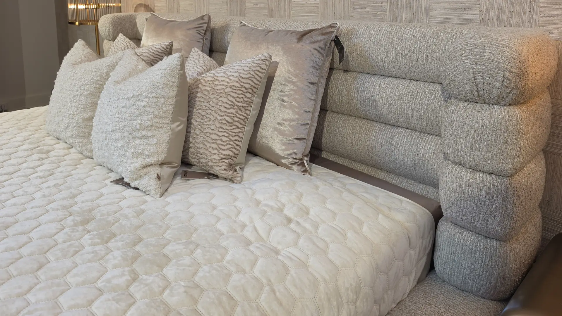 Cream quilted bedspread with several plush decorative pillows in neutral tones, set against a textured beige upholstered headboard and side lighting in a luxurious bedroom display