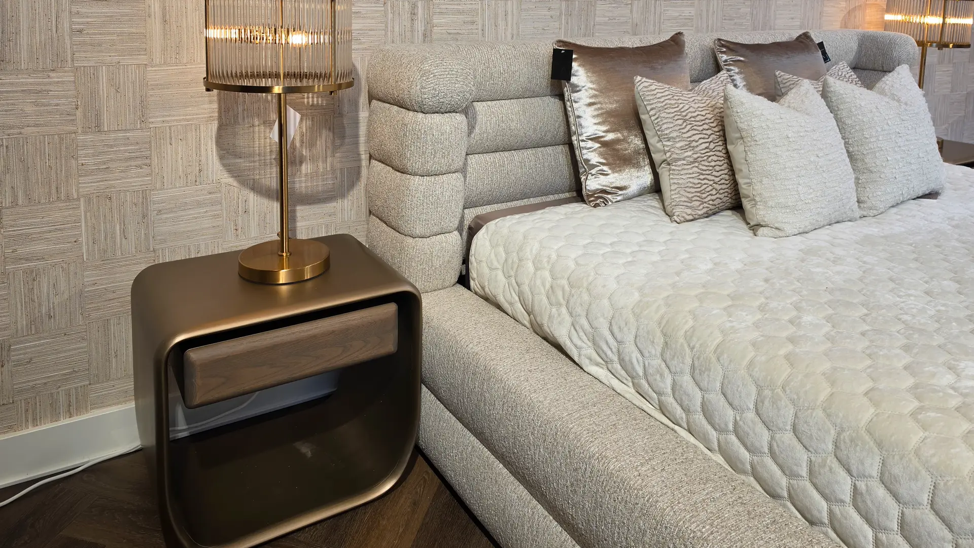 Beige upholstered bed/sofa with a quilted cream cover and decorative pillows beside a brass table lamp on a dark wood nightstand.