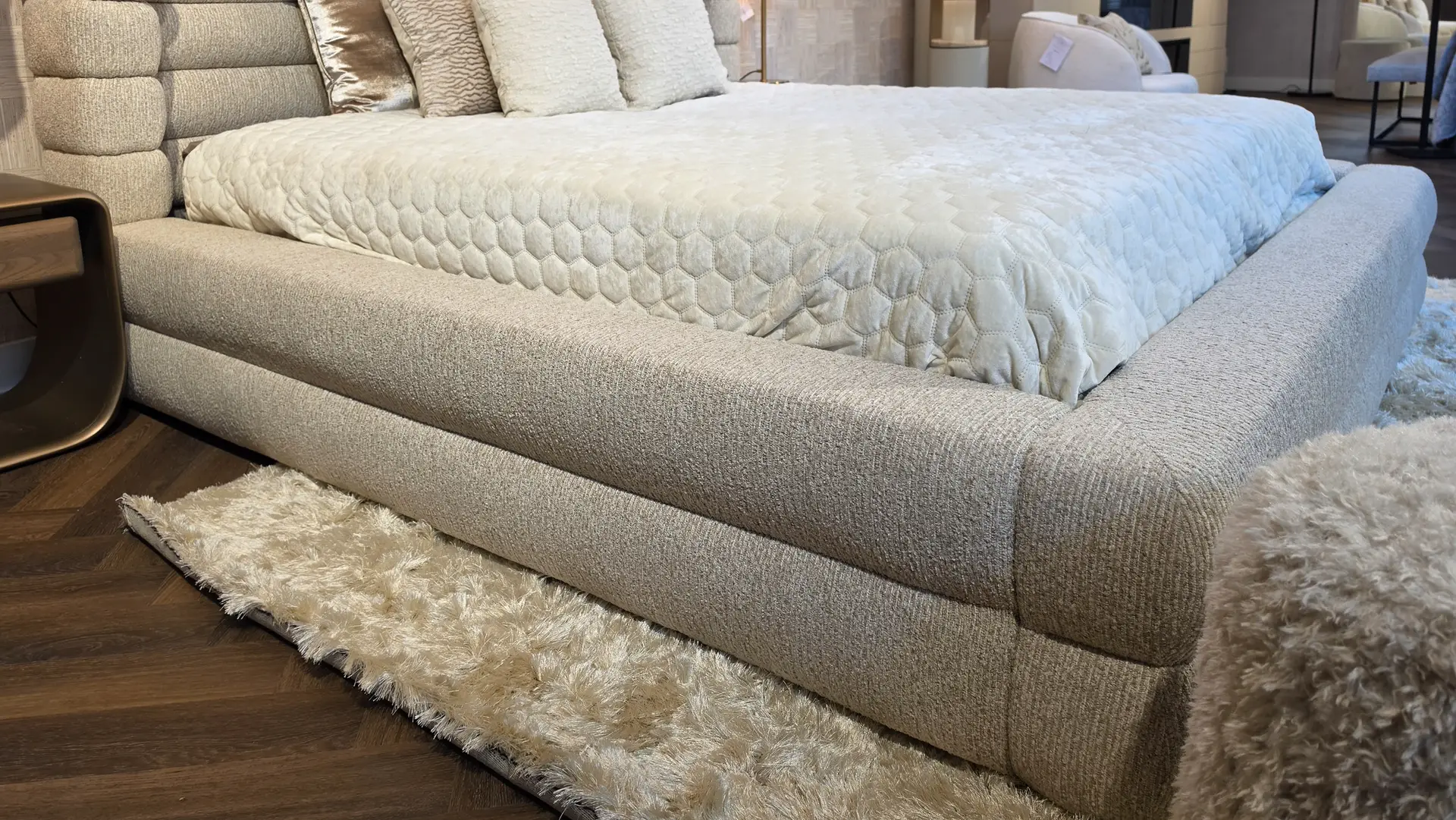Beige upholstered platform bed with a white quilted comforter and several pillows in a furniture showroom.