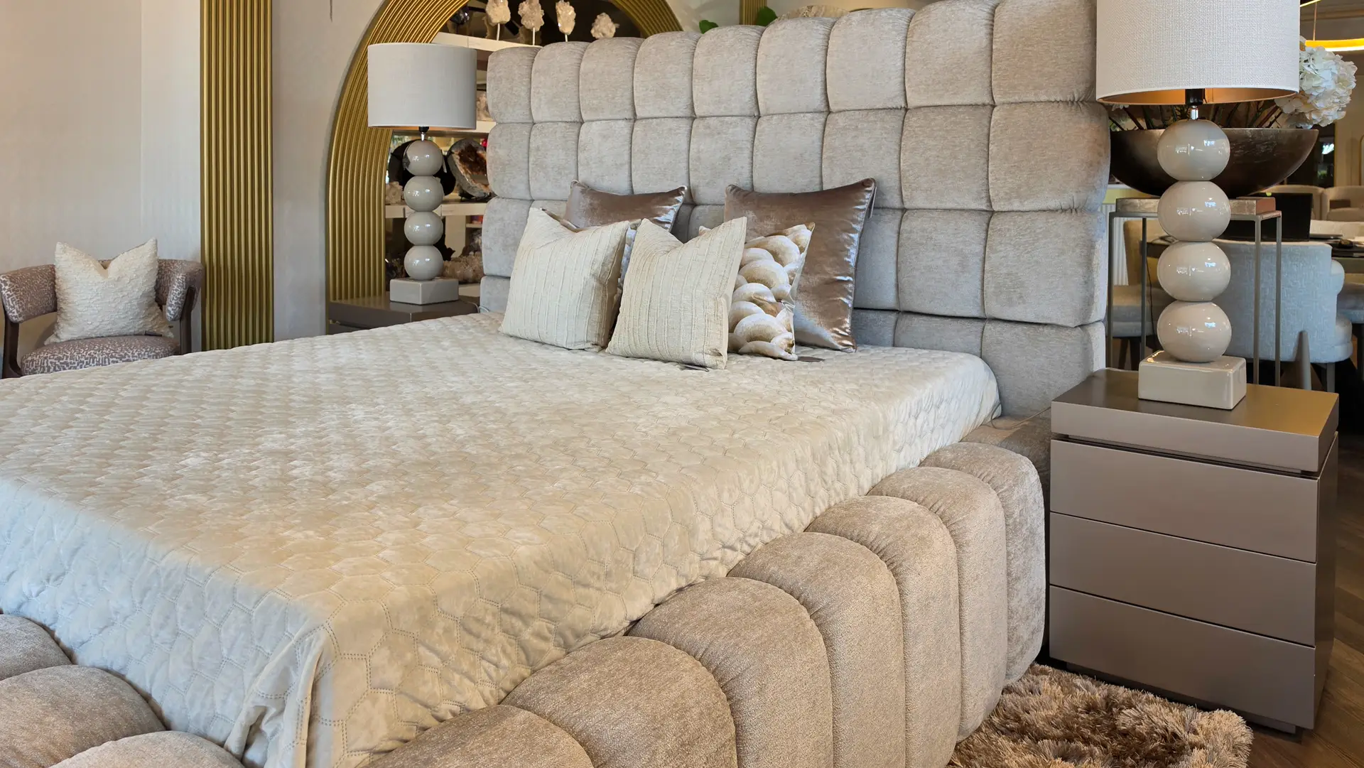 Luxurious gray upholstered bed with a tall tufted headboard, matching pillows, and two rounded-glass lamps on nightstands in a showroom-styled bedroom.