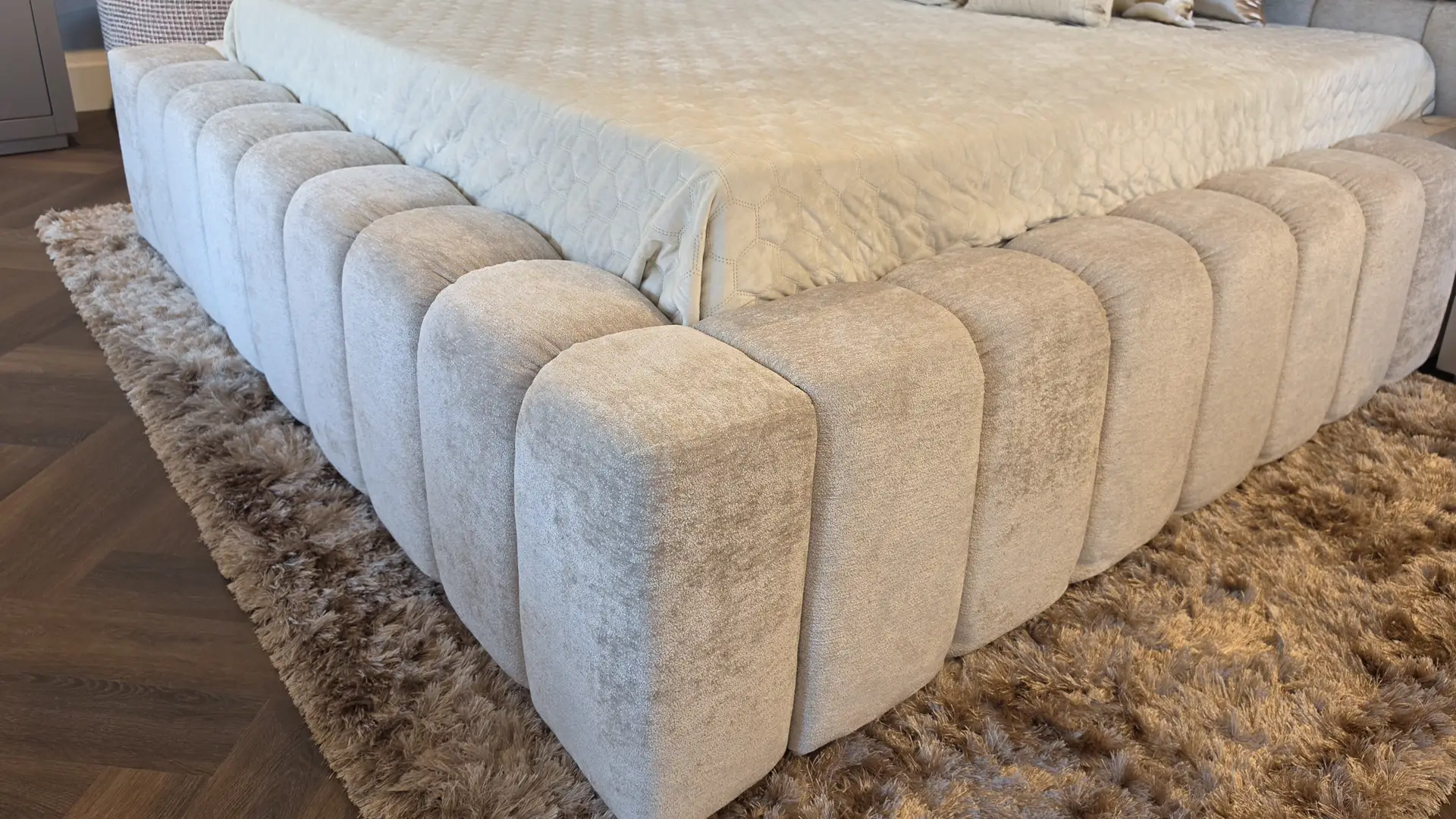 Close-up of a luxurious upholstered bed with rounded, pillow-like posts and a cream quilt on a shaggy beige rug.