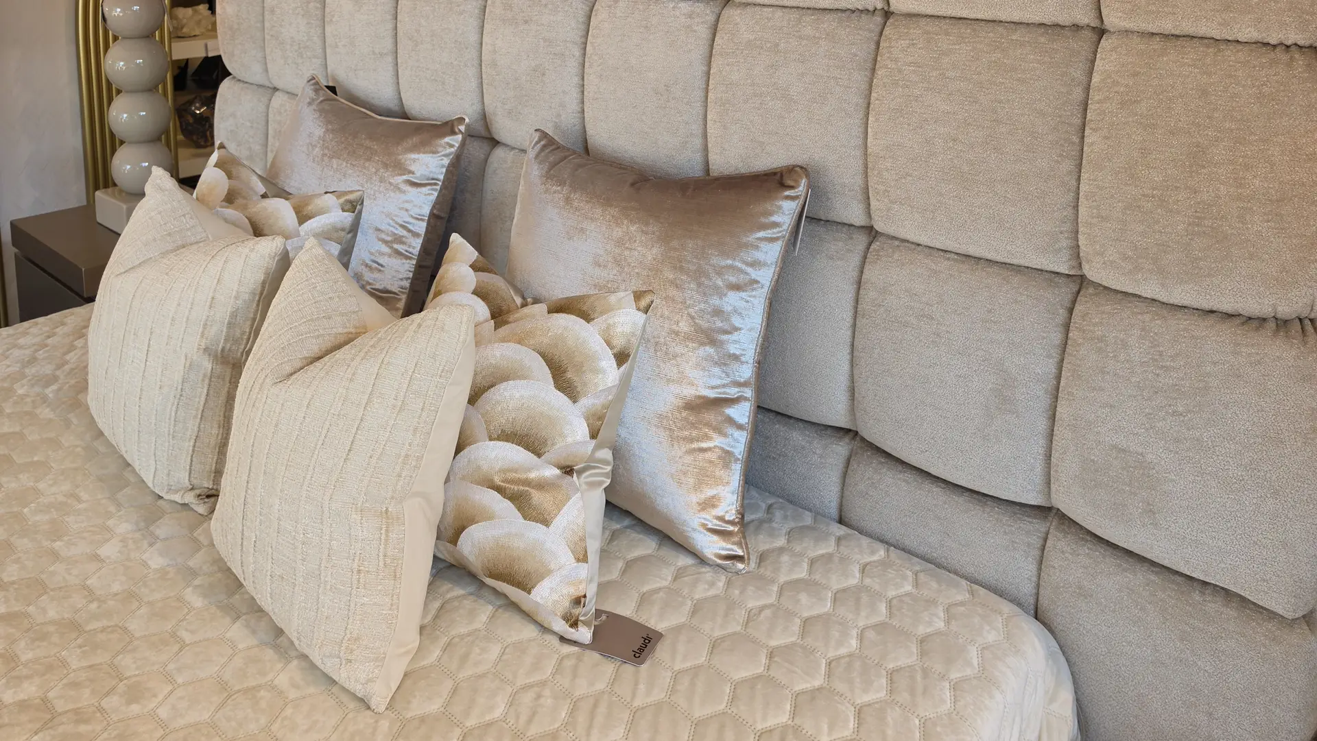 Beige tufted headboard with cream and metallic pillows arranged on a quilted bedspread.